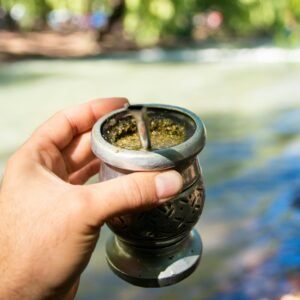 Close-up of a hand holding a traditional mate cup by a river in daylight outdoors.